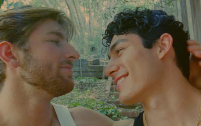 Two men in a backyard surrounded by trees and concrete blocks facing each other as one tenderly kisses the other on the forehead
