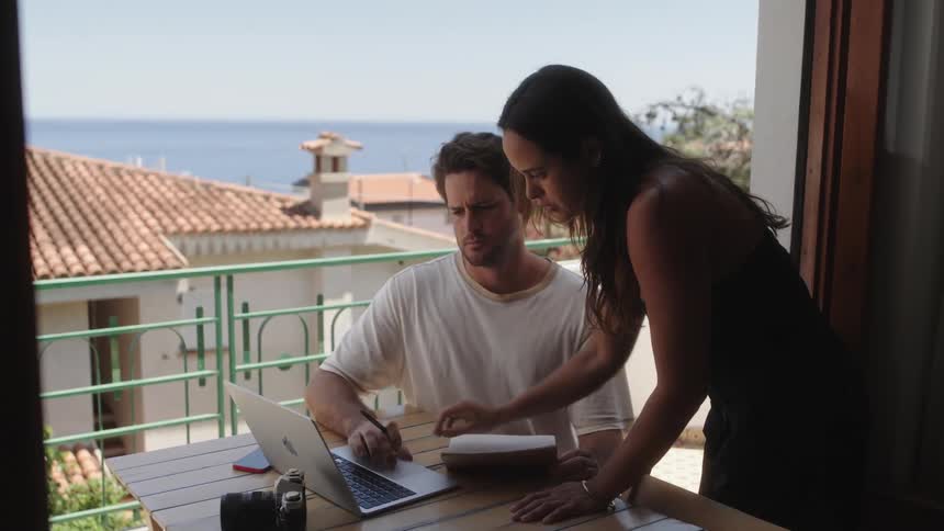 A woman holding a book while explaining something to a man seated while operating a laptop on the balcony on a sunny day