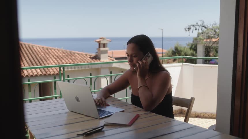 A woman sitting at a table on her balcony while talking on her cellphone and looking at her laptop on a bright sunny day