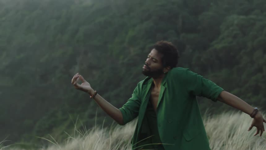 A man standing on a grassland doing hand movement with his hand while wearing a shirt without button