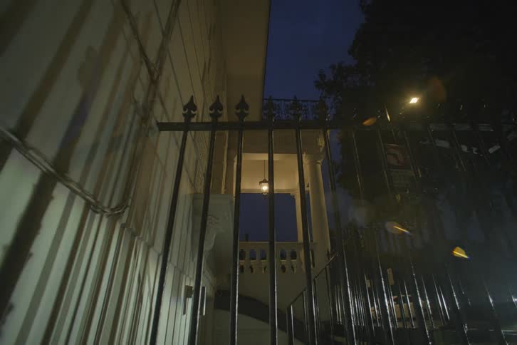 Close up of a fence gate along the exterior of a white house at night with the porch lights on
