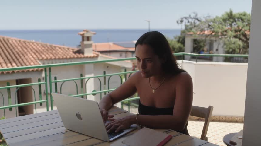 A woman sitting at a table and typing on her laptop with the sea in the background