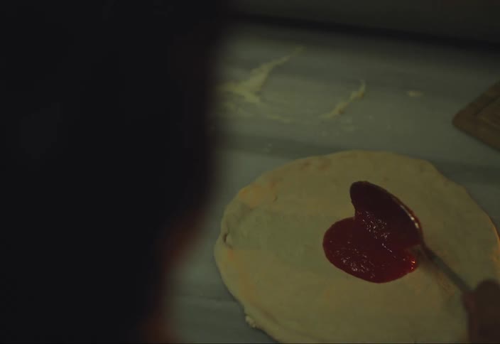 Someone uses a spoon to spread tomato sauce over dough on a countertop