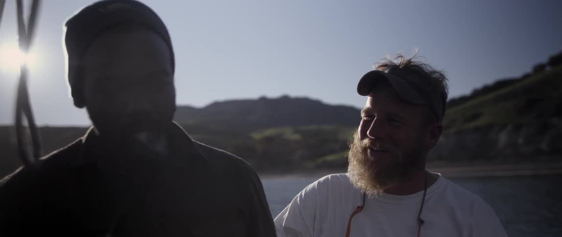 A man smokes a pipe while another man talks on a boat