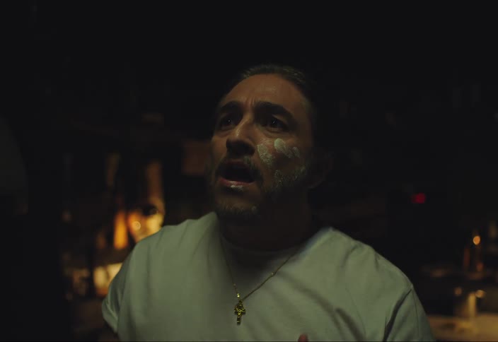A man singing in his low lit kitchen while wiping a white substance on his face