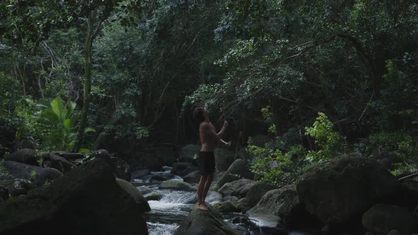 In the rainforest a barefoot man wearing shorts jumped onto a rock in a flowing stream holding onto a rope