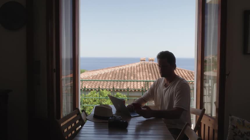 A man sitting in front of a window working on a cell phone and a notebook