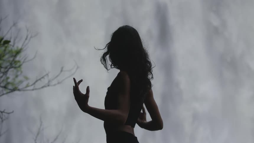 A woman standing in nature in front of a water fall while she stretches her arms out to the sides doing yoga