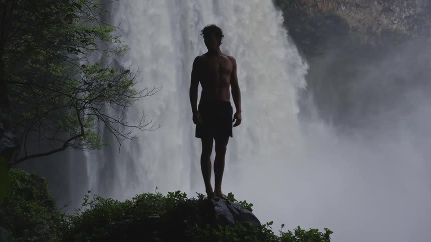 A man is perched on a stone by the waterfall