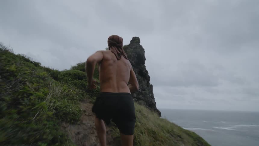 A shirtless man running on a trail of vegetation and on the right side there is a slope to the ocean