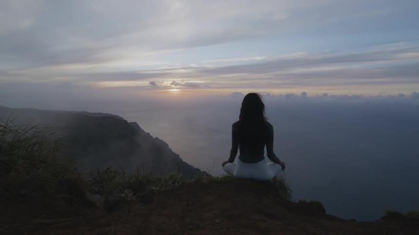 Woman sits to meditate at the edge of a cliff next to the ocean at sunset