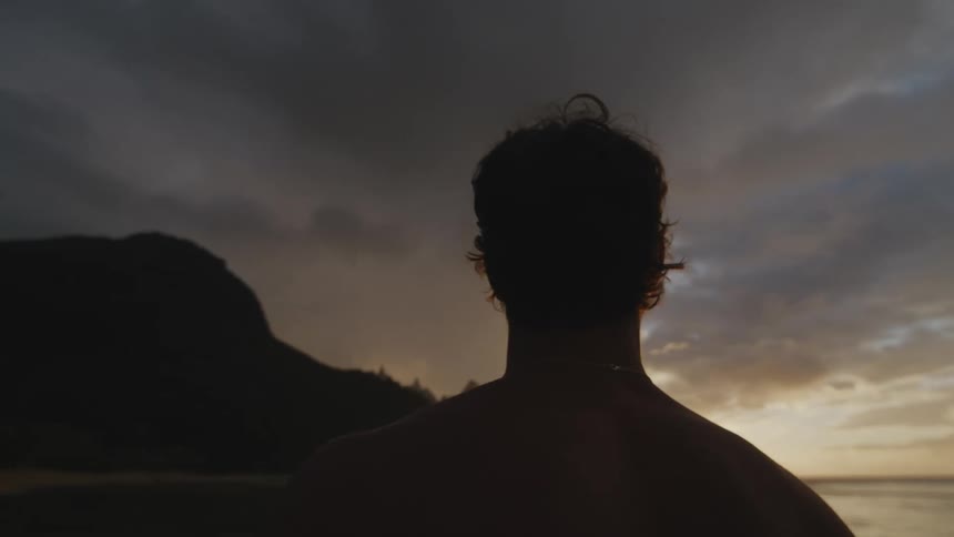 A shirtless man stands on a beach at dusk taking in his surroundings
