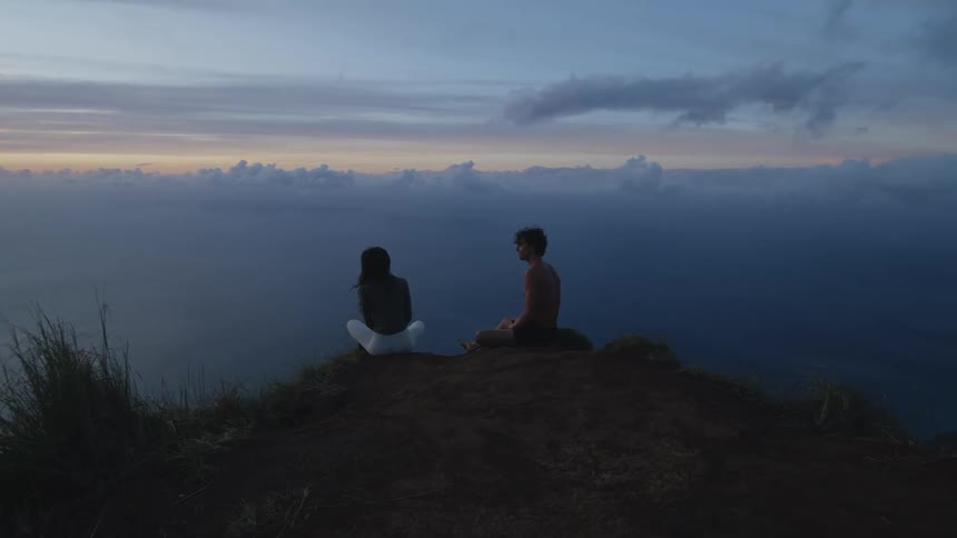 A man and a woman seating at the edge of a cliff looking at the cloud with fog covering the atmosphere in the evening