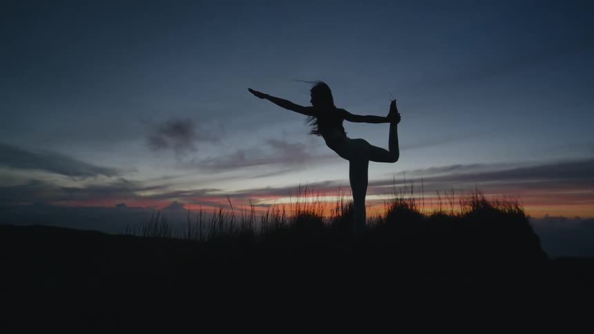 A silhouette of a woman stretching and doing yoga in a grassy field as the sun sets on the horizon behind her