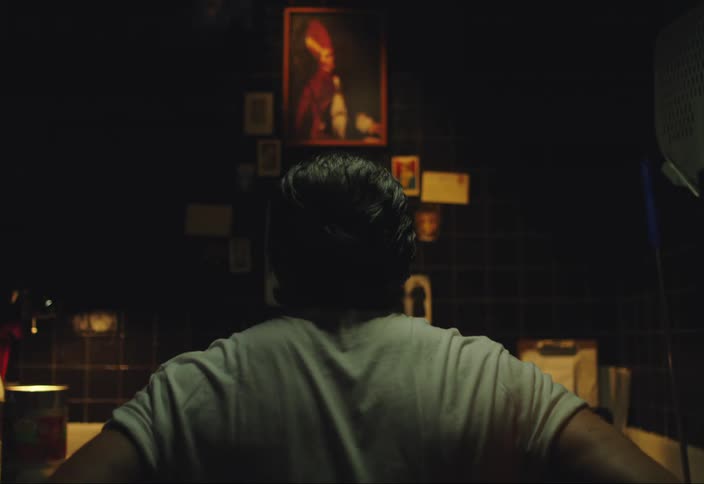 A man sits looking at an altar with a painting of a person hanging on the wall