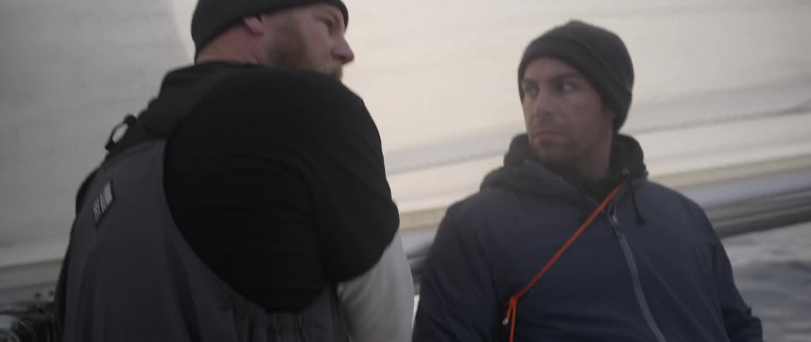 A man is talking with another man pointing up at a sail on a boat
