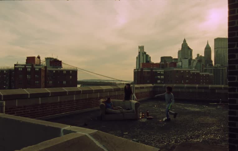 Three children run and sit on a couch on top of a highrise building in a city at sunset