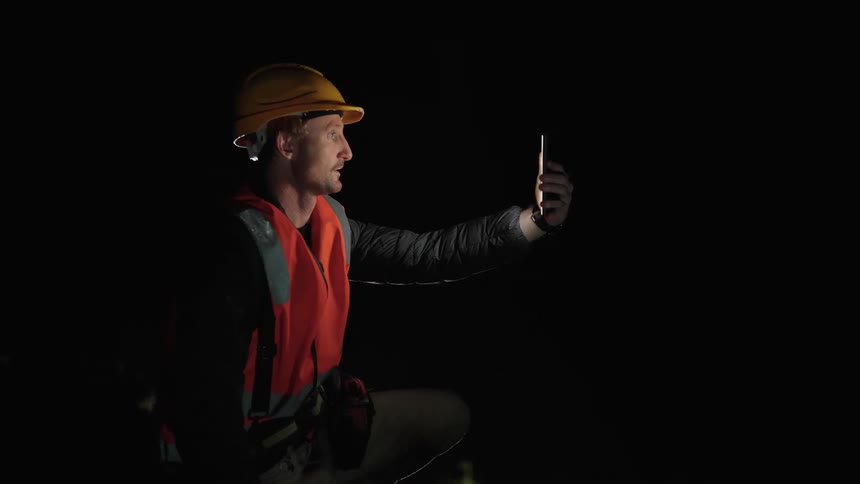 A man wearing a helmet safety vest and tool belt kneels while holding a cell phone flashlight and talking to someone at night