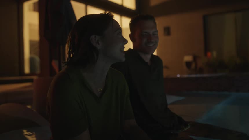 A man and woman sit and laugh together outside with their legs in a pool next to a building at night