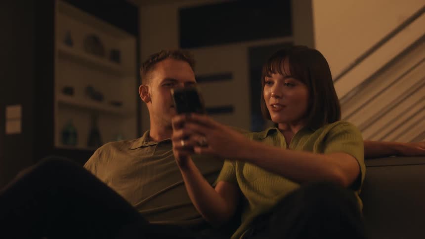 A man sitting on the couch then a woman sits next to him using her cellphone and both converse with smiles