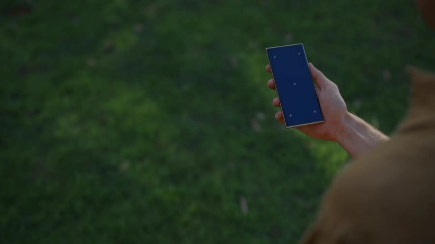 A man walks on a lawn holding a cell phone that shows a dark screen with 5 white dots
