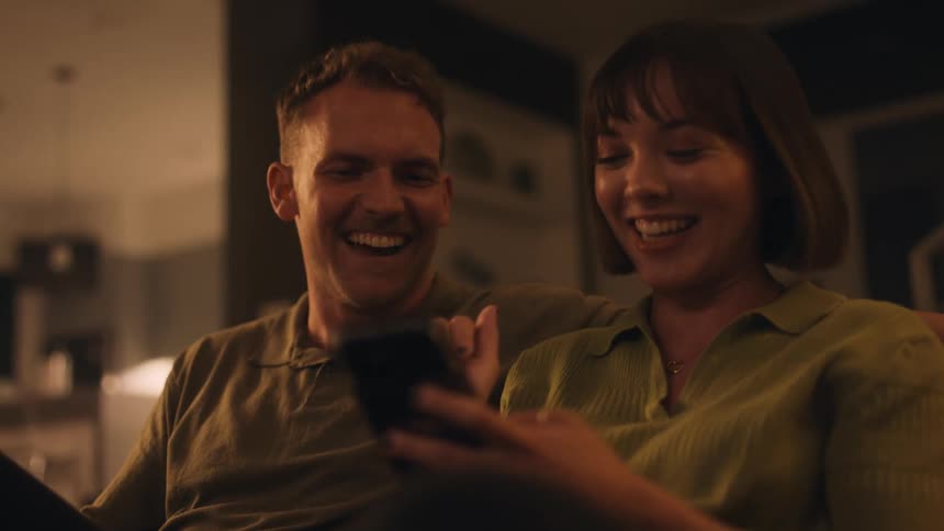 Man and woman talking and smiling while looking at a mobile phone sitting on a couch indoor