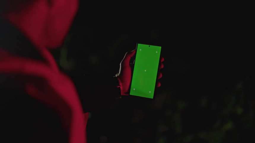 A man illuminated in blinking red light looks at a phone that shows a green screen and white dots outside at night