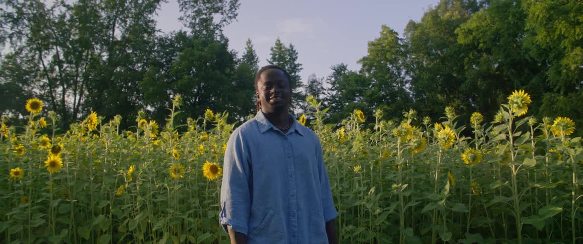 A black man with dreadlocks wearing a blue sirt standing in a farm with yellow flowers
