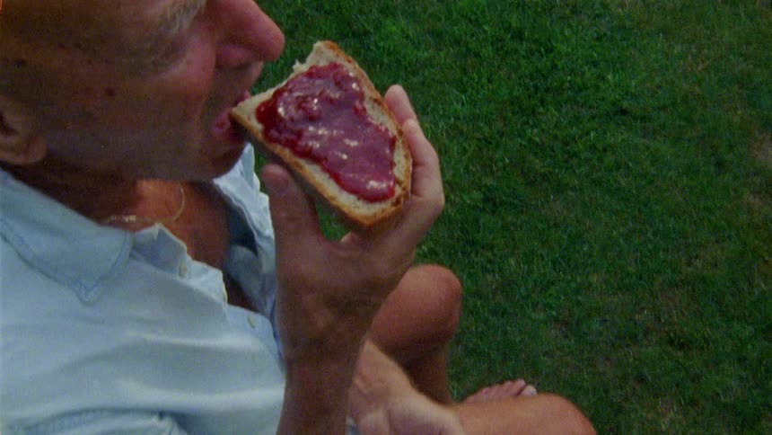 Elderly person sitting in a backyard preparing a bread with jam and then starts eating
