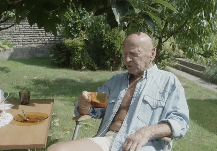 An old man sitting and drinking tea with his buttondown shirt open