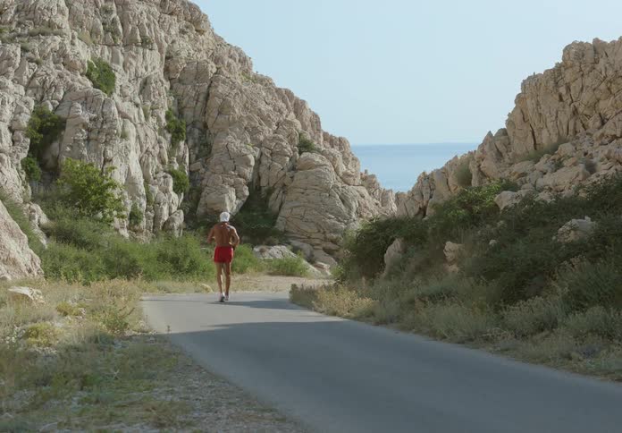 Shirtless man with a white cap jogging alone in day time on a road inbetween rocks