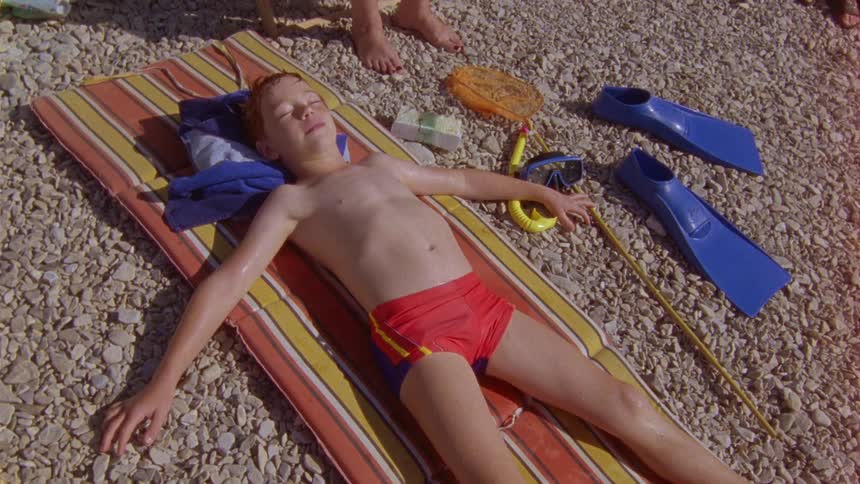 A boy lays on a raft in his swim shorts atop a stonecovered ground