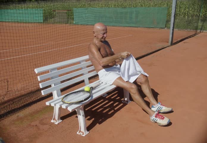 An elderly person on a tennis court sweaty leaves his racket on the bench and sits down he then dries his face with a towel and places it on his face