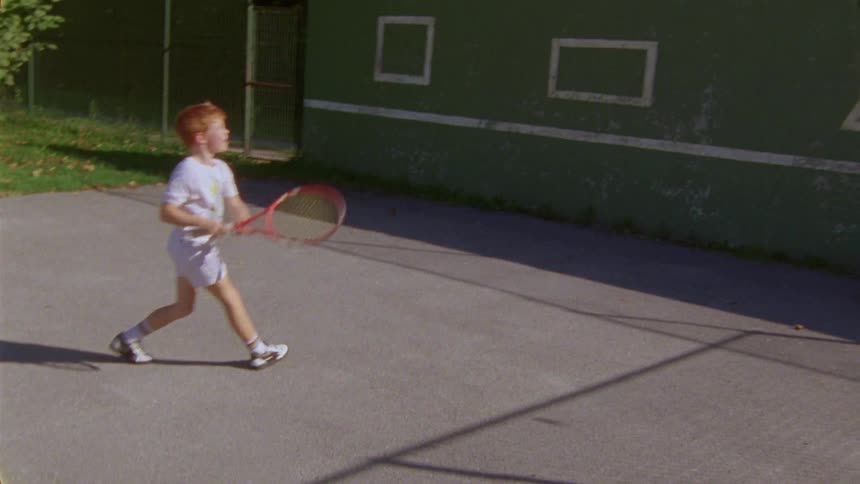 A boy hits a tennis ball with a racket against a wall with white shapes painted on it