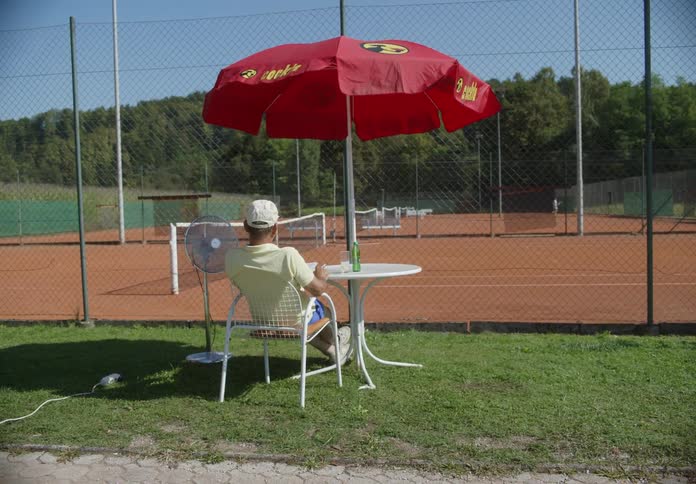 A man sits at a table with an umbrella while watching someone play tennis in a court