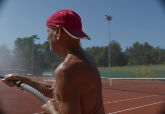 An older man wearing a red baseball cap backwards using a hose to spray down a tennis court on a bright sunny day