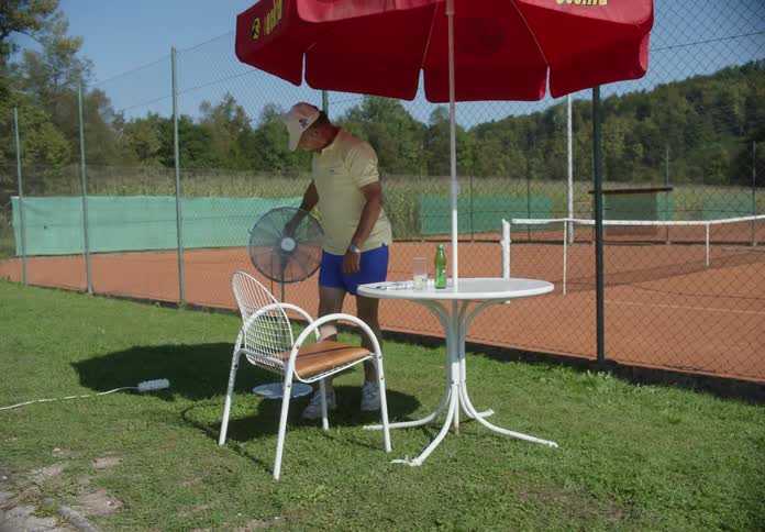 A man switches on a standing fan then settles into a chair beneath an umbrella at a table near the tennis court