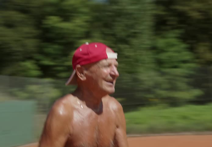 An elderly man plays tennis on a court on a hot summer day