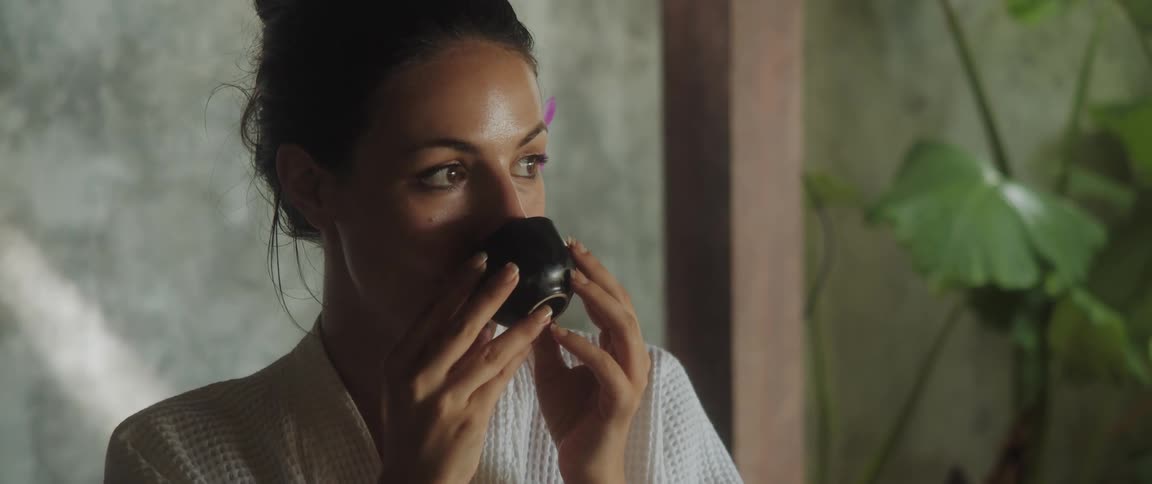 A woman with a flower in her ear sipping something from a cup while pensive