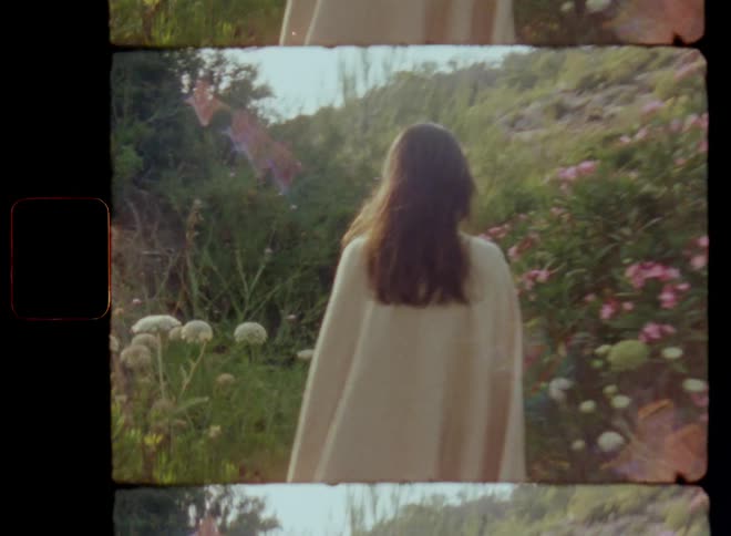 Woman with long brown hair wearing a yellow towel or blanket as a cape walks through flowering wildflower bushes on a hidden path or way in a small valley in a hilly countryside in the morning time enjoying a stroll in nature