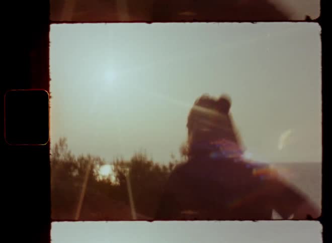 Handheld dynamic 8mm film footage with shaky camera movement of sportive young man who jogs uphill along trail overlooking ocean on sunny day as his dark silhouette moves towards bright sun shine causing lens flare and glare with stunning sea view