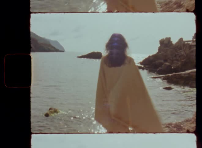 Vintage Super 8 camera shot following brunette woman with long loose hair wearing a yellow blanket over her shoulders swaying or moving in the wind walking to the shore of the sea with several stones in the water in a hidden bay with beautiful beach