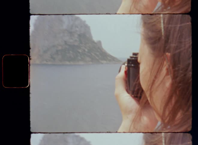 Brunette woman raises or lifts old vintage SLR camera with her hands up to her eye to take photo of a rock formation located near the coast to capture her sightseeing experience via analog photography as a tourist on touristic island on Super 8mm film