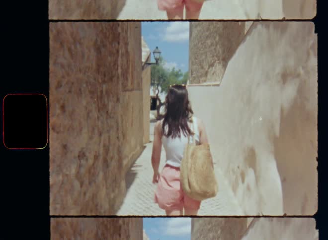Backside of a brunette woman wearing summer clothing walking down a sidewalk or path in a fast way between buildings and stone walls on a bright sunny day towards a tree standing on a market square of an old historical city in the south