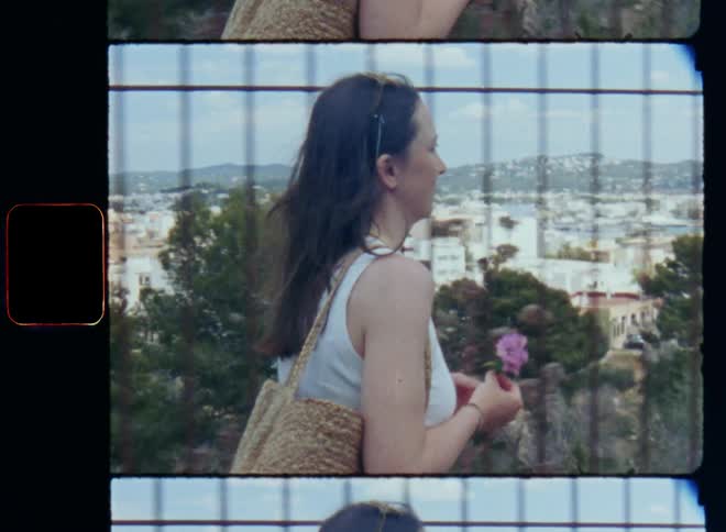 Young lady carrying a handbag and a small flower bouquet in her hands walking by the sidewalk or pavement on a hill in front of a fence surrounding a garden with green bushes and trees with a cityscape or town in the valley in the background
