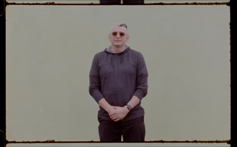 A man standing in front of a white wall outside while wearing sunglasses and his hands together in front of him