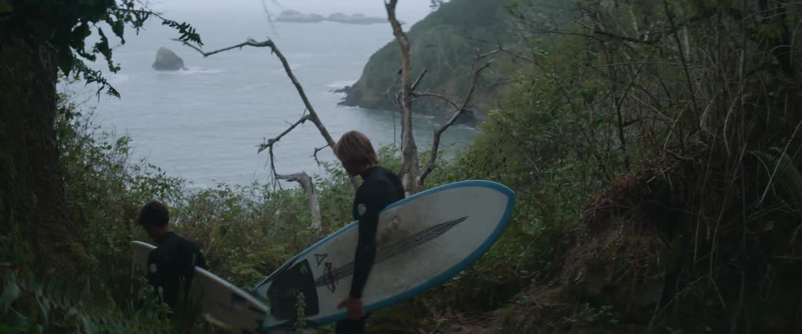 Two young men holding their surf board stepping out of the jungle heading into the sea to surf