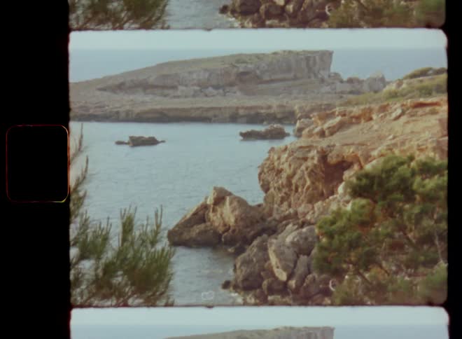 Landscape Super-8 film footage with sidewise horizontal camera movement of a coastal region of stones and rocky cliffs on the coast of the sea or ocean with plants and vegetation and islands blocking the view to the horizon on a sunny day