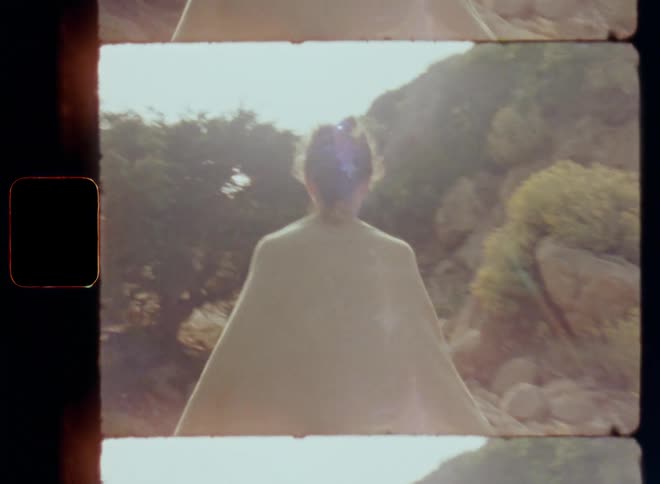 Brunette lady with chignon or bun hairstyle and a bathing towel walking alone on a path near big rocks approaching a hidden beach in an idyllic bay at the sea ocean or lake on a sunny day on vintage Super-8 film with bright lens flare