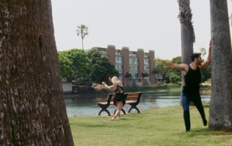 Two dancers dance in park across water from apartment building
